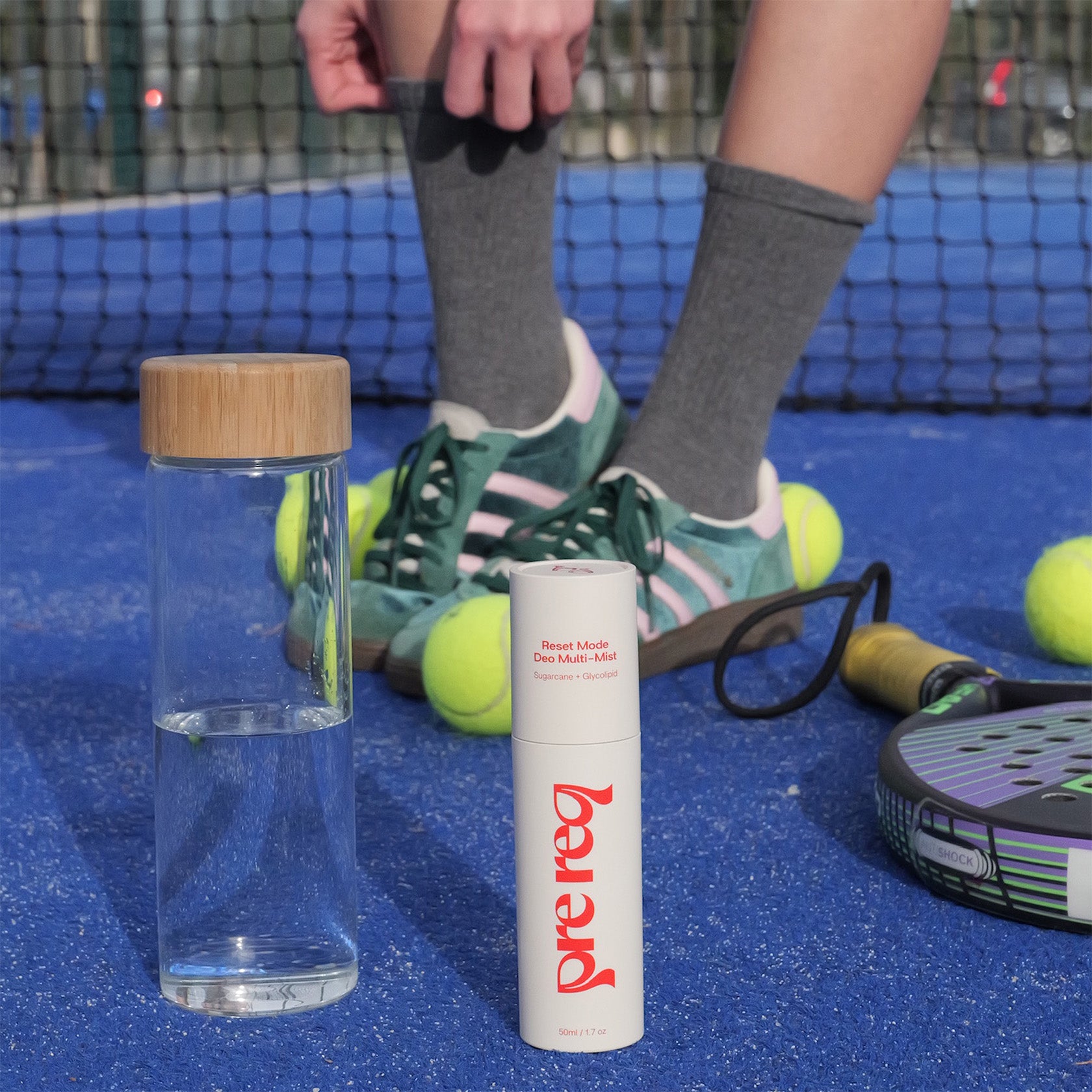 Clear water bottle with wooden cap and white Prereq supplement container on a tennis court.