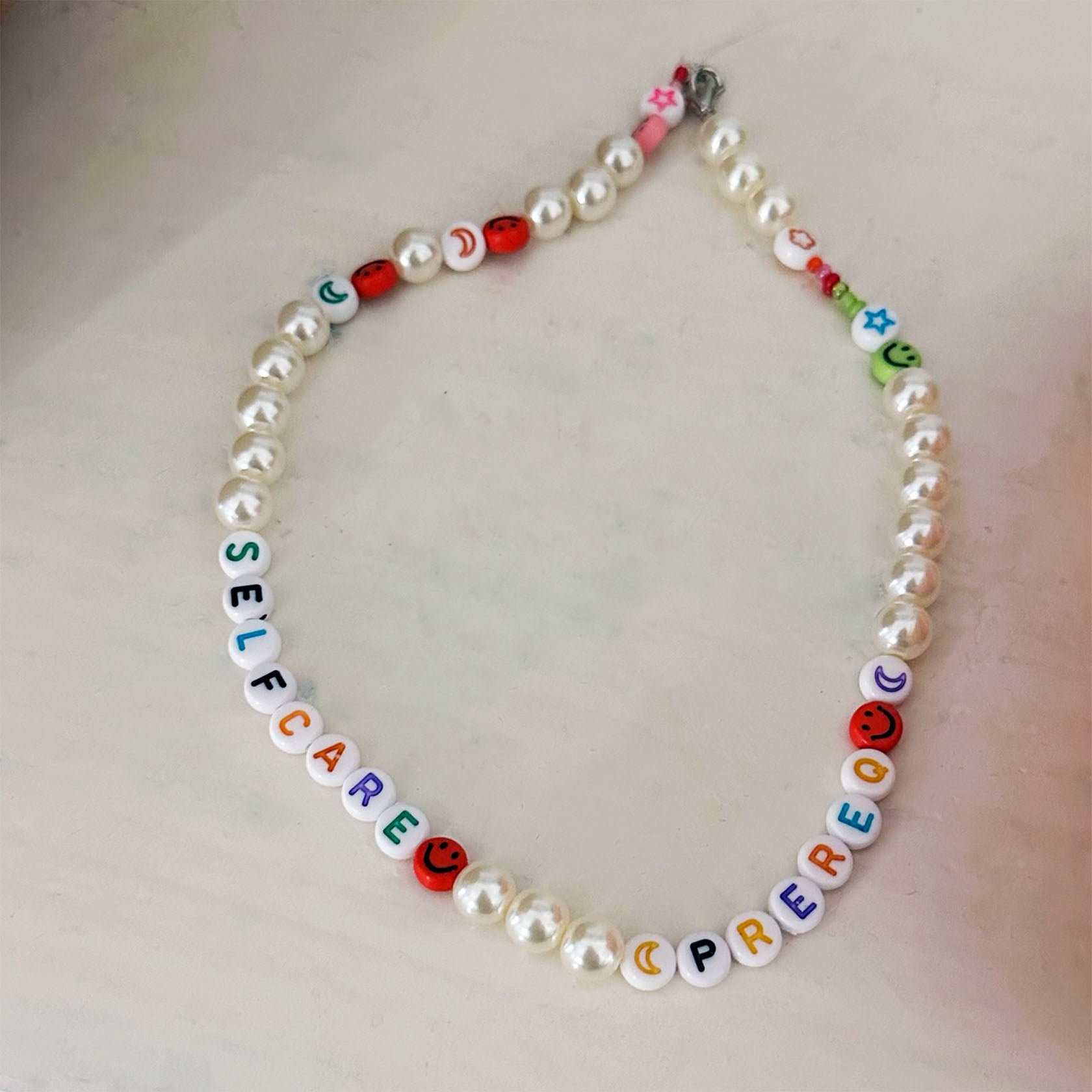 Necklace with letter beads spelling 'SELF CARE' and colorful emoji beads on a light background
